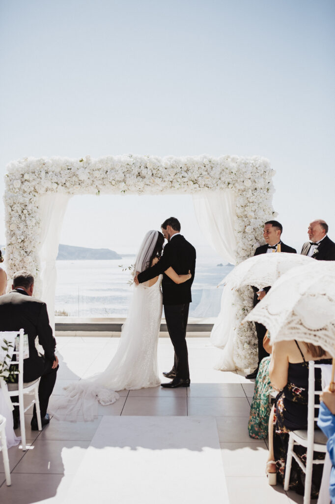 real radio and groom hug each other at an outdoor ceremony at le Ciel Santorini