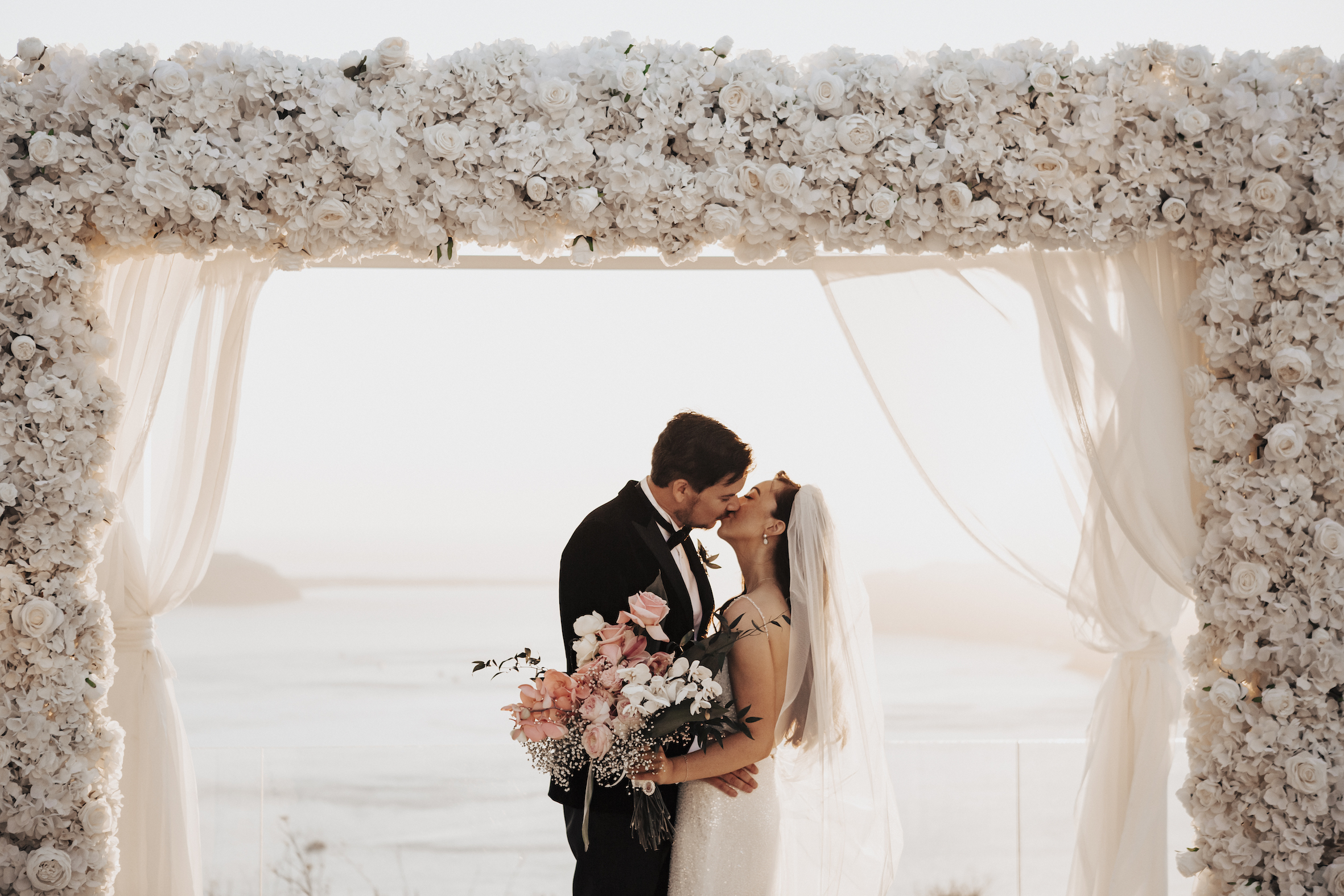 Anneliese and James kiss under white floral arch at Le Ciel wedding Santorini