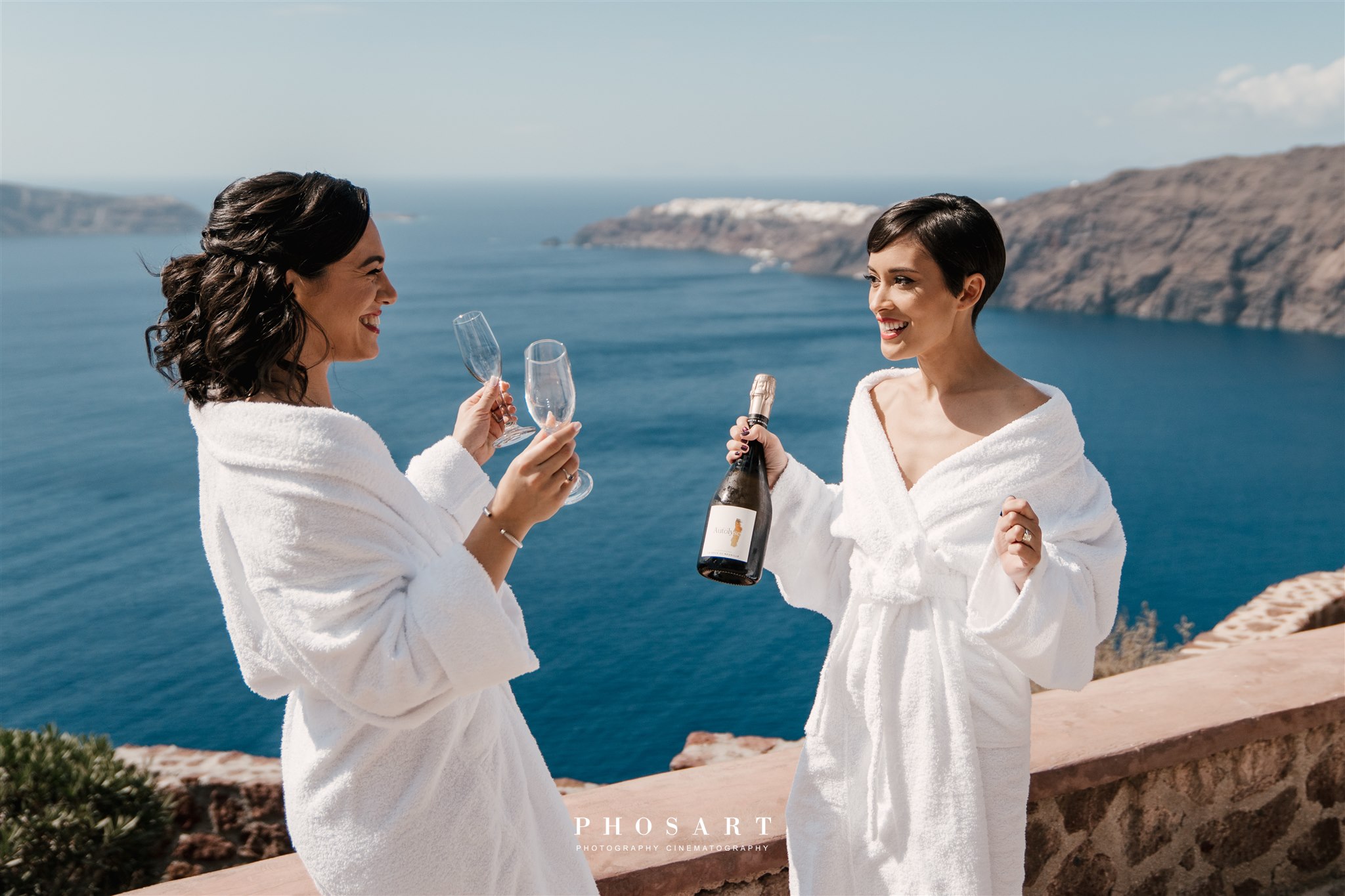 Bride and bridesmaid holding champagne bottle and glasses at Le Ciel Santorini wedding venue