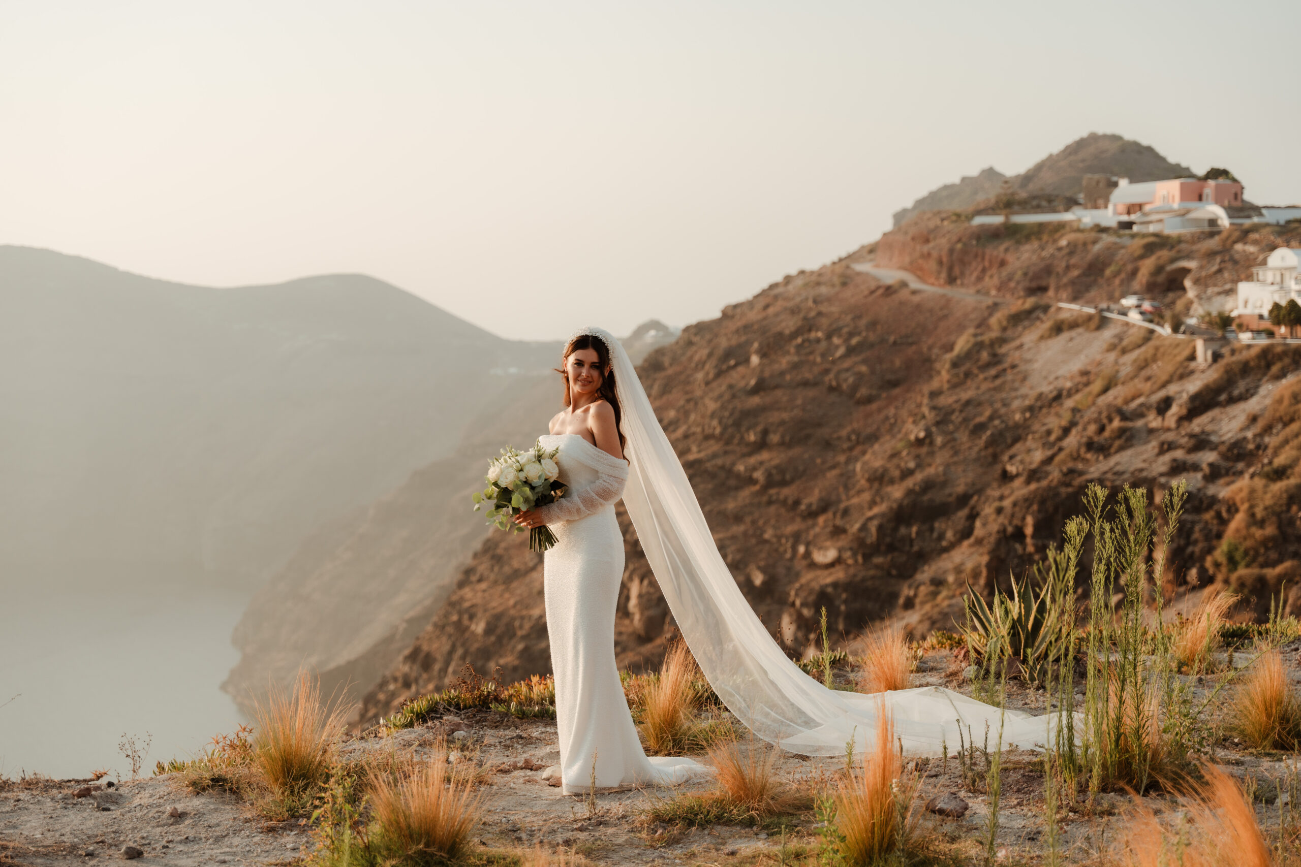 bride standing on clifftops of Santorini at destination wedding wearing dress with tulle sleeves