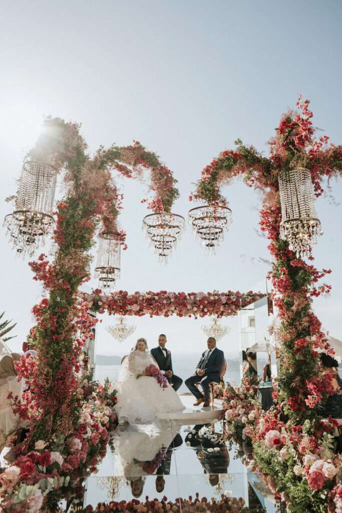 Leeda and Varun bride and groom sitting underneath big floral display at Le Ciel wedding